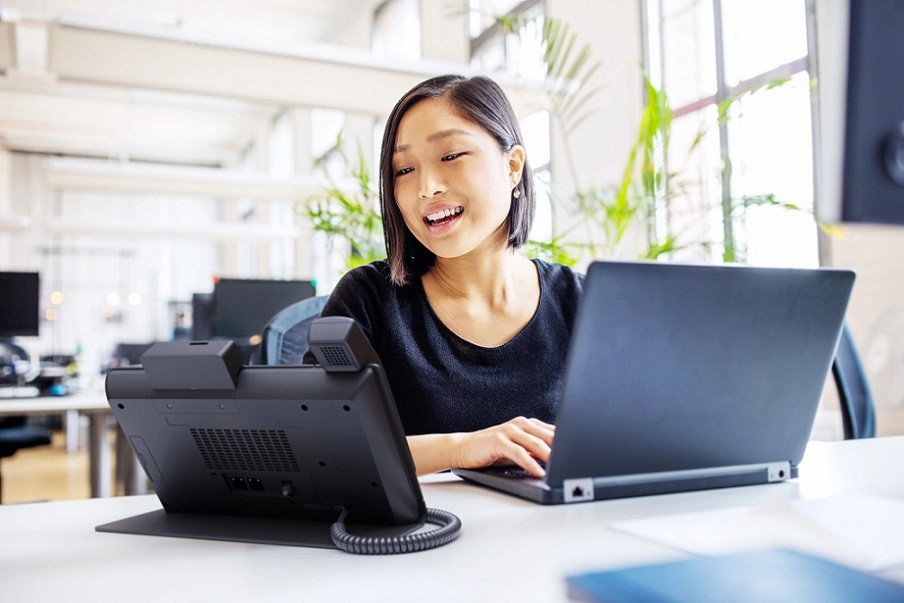 An employee speaking on a Crestron Flex phone while sitting at her desk typing on her laptop.
